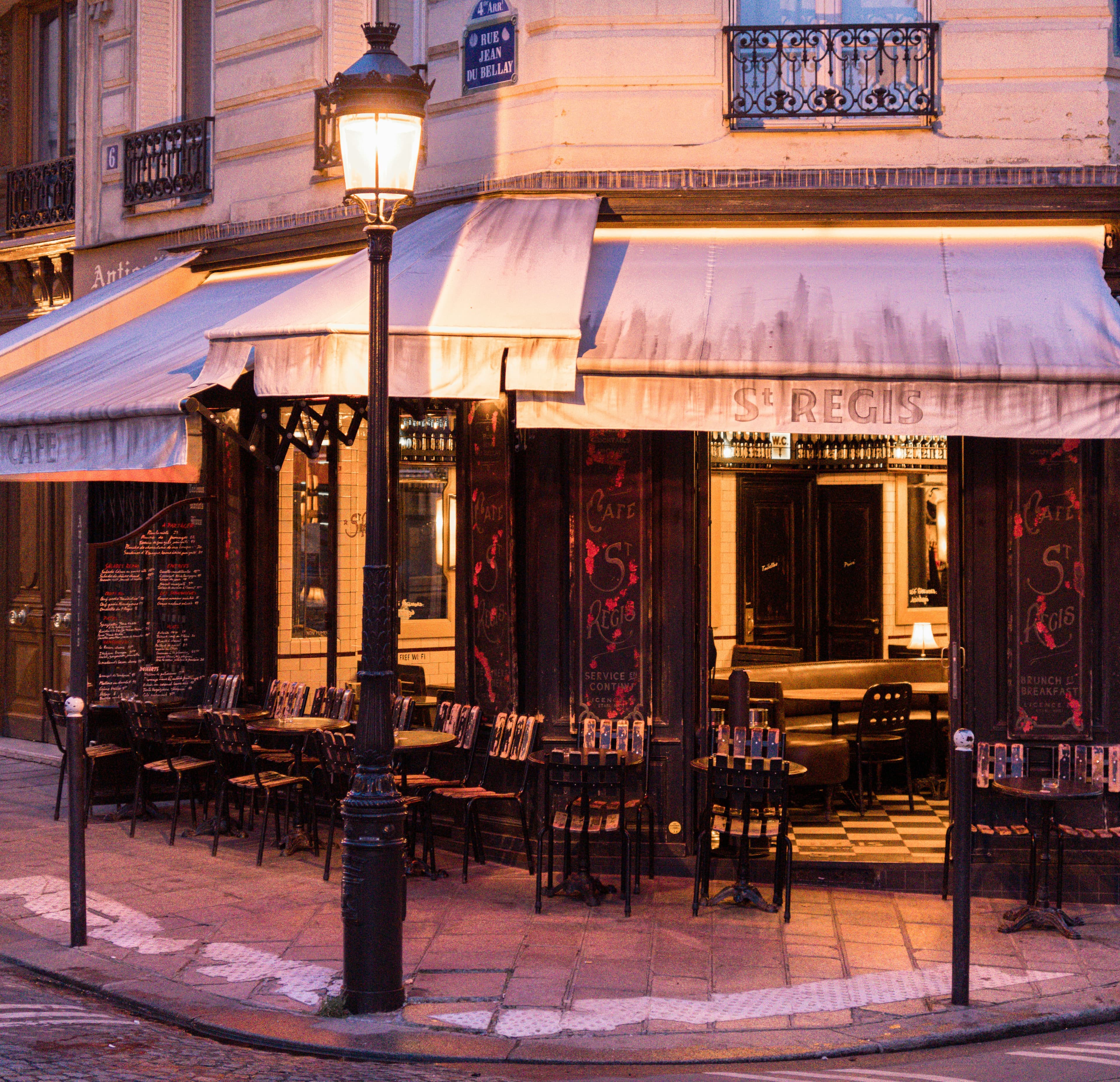 A corner café in Paris at dusk, its awnings and pavement tables lit by a street lamp